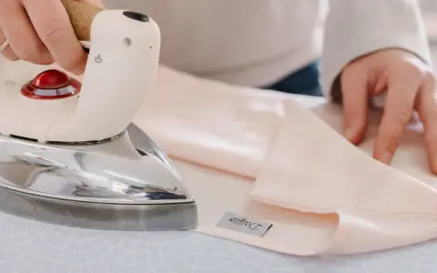 Mani che stirano lenzuola rosa pallido con un ferro da stiro Immetec. Un'etichetta con il logo "Daffodil" è visibile sul tessuto.