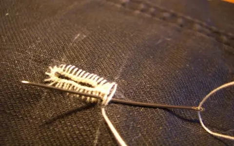 Ago e filo bianco creano un punto occhiello a mano su tessuto scuro.