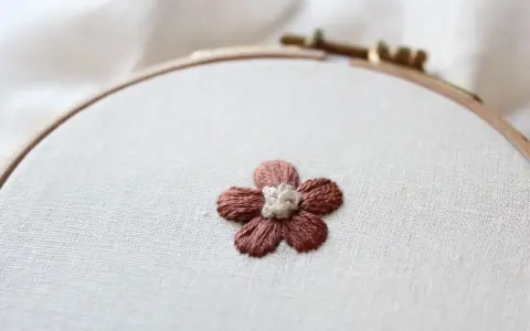 Piccolo fiore ricamato a punto pieno su tela grezza, con petali marroni e centro bianco.