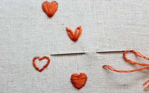 Un ago con filo arancione pronto per creare un altro cuore ricamato a mano su tela grezza.