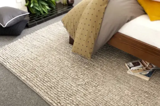 Un grande tappeto di lana intrecciata accanto a un letto. Scopri come lavare un tappeto di lana grande per mantenerlo come nuovo.