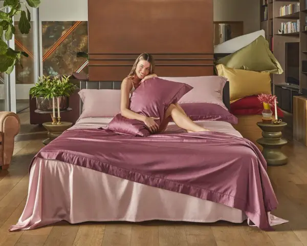Donna sorridente su un letto con lenzuola rosa e copriletto prugna, che illustra la differenza tra piumone e trapunta.