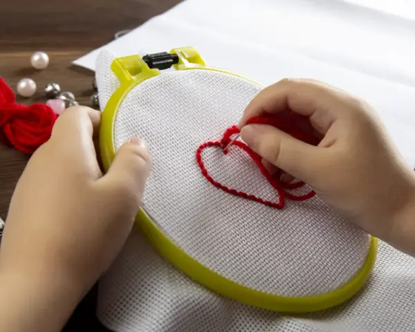 Mani di bambino creano un cuore rosso con il punto erba a mano su tela bianca, circondato da perle e gomitoli di lana.