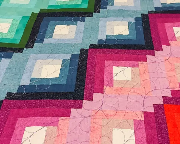 Dettaglio di una coperta realizzata con la tecnica patchwork, con blocchi a gradiente di colore dal blu al rosa, rifinita con cuciture decorative.