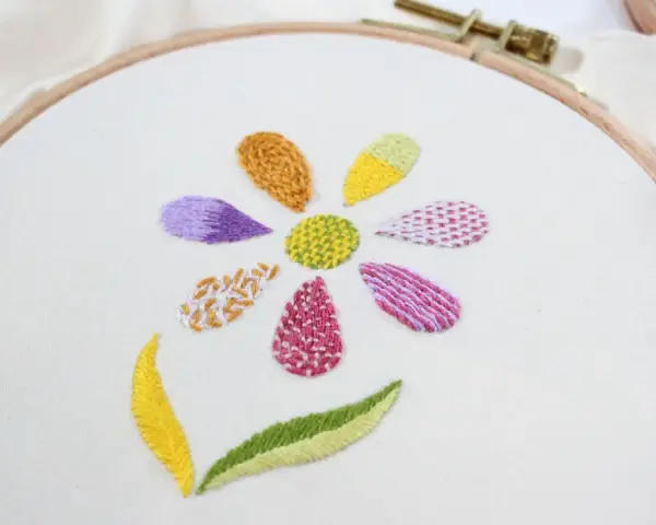 Fiore ricamato su tela bianca con petali colorati e diversi punti ricamo per riempire, che mostrano varie texture e tecniche.