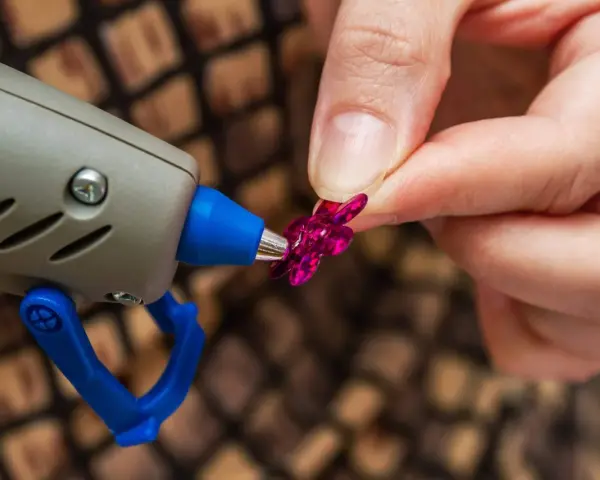 Applicazione strass su tessuto con colla a caldo. Una mano tiene un fiorellino fucsia mentre la pistola a caldo è pronta per incollare.