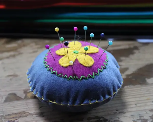 Puntaspilli fai da te a forma di fiore, con petali gialli su sfondo viola e base blu, decorato con cuciture a zig-zag verdi e gialle.