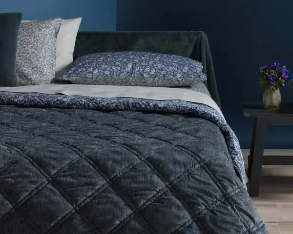 Letto con copriletto trapuntato blu scuro, cuscini floreali e un dettaglio che ricorda che il velluto si rovina con acqua.