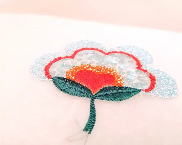 Un fiore stilizzato con petali azzurri e paillettes, un cuore rosso al centro e perline arancioni. Un'opera di ricamo francese che cattura la luce.