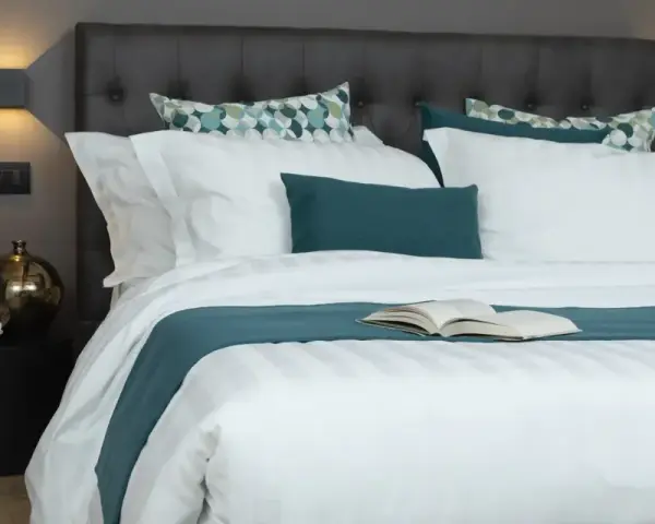 Letto rifatto con lenzuola bianche e copriletto turchese. Un libro aperto è posato al centro, pronto per essere letto. Ecco come si mette il lenzuolo di sopra.