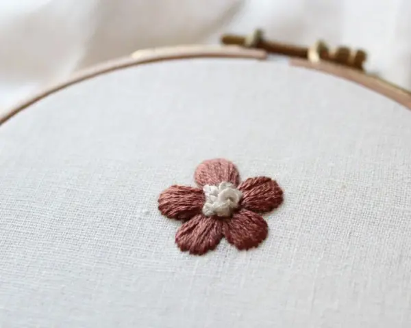 Piccolo fiore ricamato a punto pieno su tela grezza, con petali marroni e centro bianco.