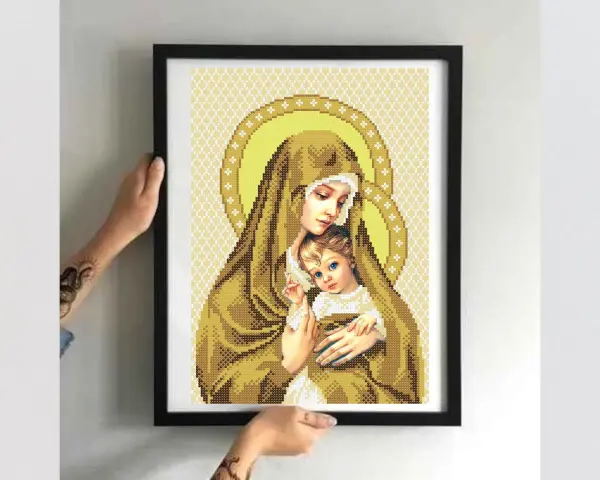 Cross-stitch of Madonna and Child in a black frame, held by hands.