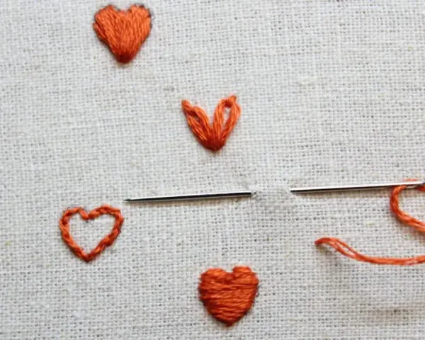 Un ago con filo arancione pronto per creare un altro cuore ricamato a mano su tela grezza.
