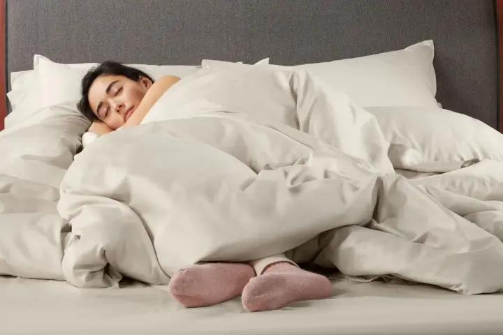 Donna dorme avvolta in lenzuola in microfibra che sembrano farla sudare, con solo i piedi in calzini rosa visibili.