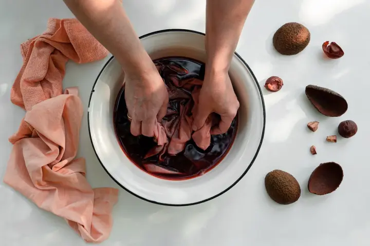 Mani che tingono tessuti in una bacinella con liquido scuro, circondata da bucce di avocado. Un processo naturale per come tingere i tessuti.