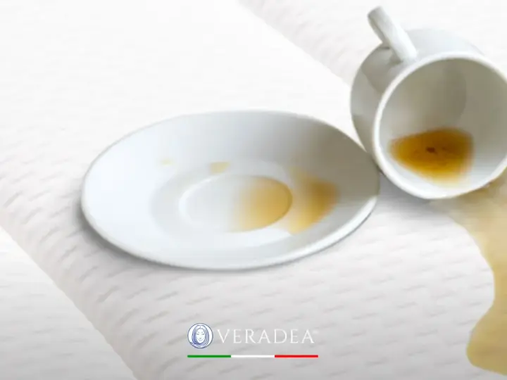 Tazza rovesciata su un materasso bianco, con liquido marrone versato. Scopri come disinfettare il materasso per eliminare macchie e odori.