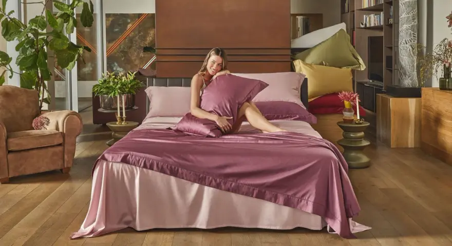 Donna sorridente su un letto con lenzuola rosa e copriletto prugna, che illustra la differenza tra piumone e trapunta.