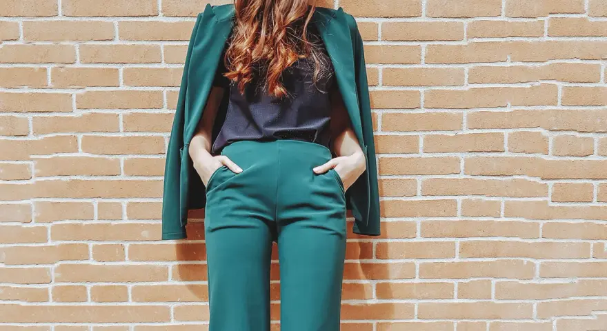 Donna con tailleur verde smeraldo, composto da giacca e pantaloni a palazzo, con le mani in tasca, appoggiata a un muro di mattoni.