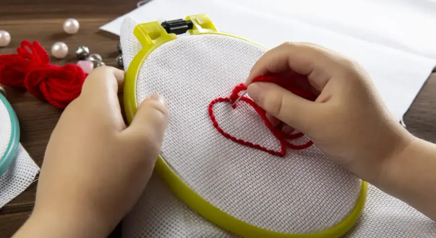 Mani di bambino creano un cuore rosso con il punto erba a mano su tela bianca, circondato da perle e gomitoli di lana.