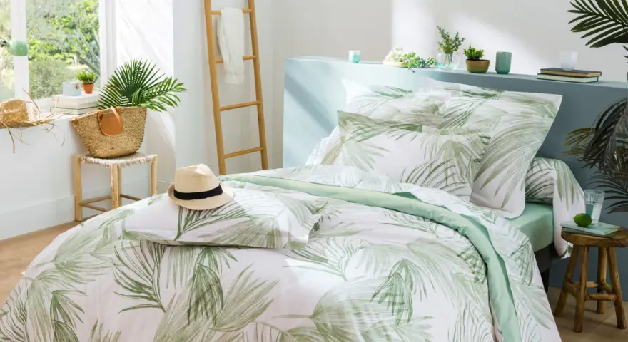 Un letto preparato per l'estate con biancheria a tema tropicale, un cappello di paglia e una borsa di vimini.