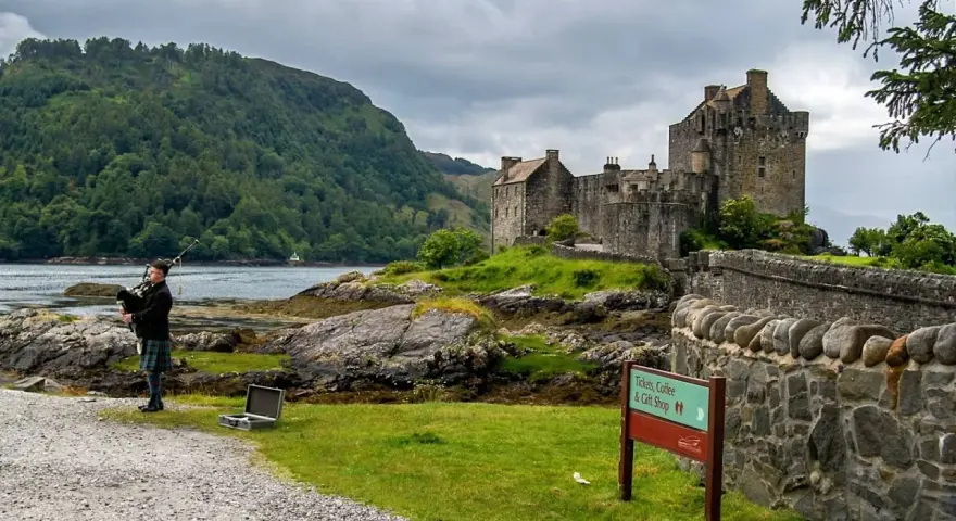 Uomo con cornamusa indossa un kilt scozzese davanti a un castello.