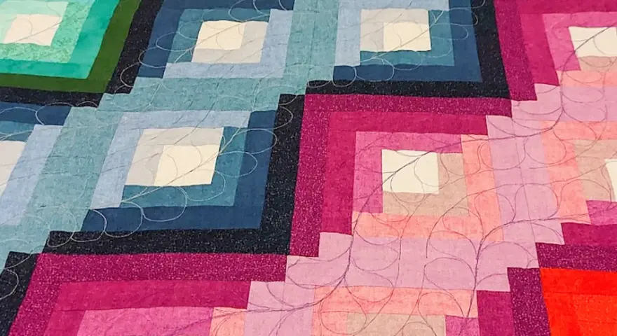 Dettaglio di una coperta realizzata con la tecnica patchwork, con blocchi a gradiente di colore dal blu al rosa, rifinita con cuciture decorative.