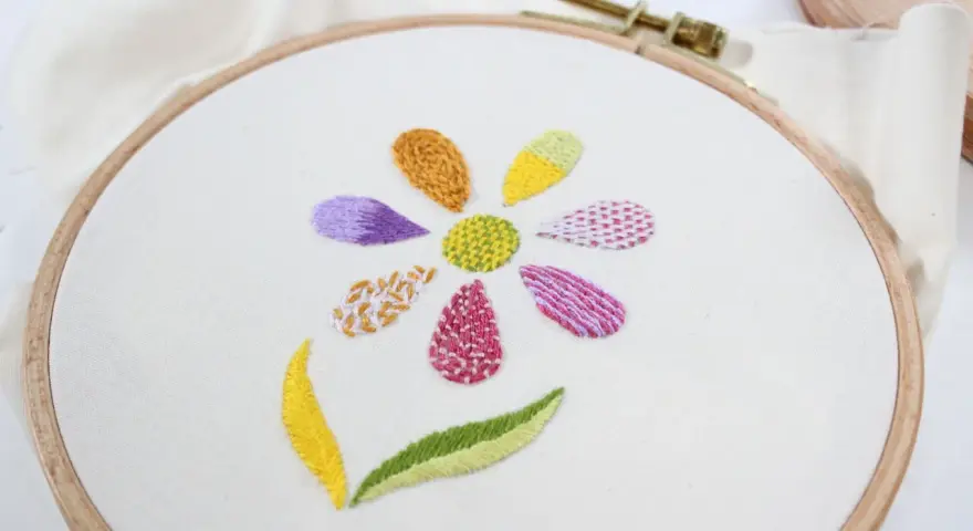 Fiore ricamato su tela bianca con petali colorati e diversi punti ricamo per riempire, che mostrano varie texture e tecniche.