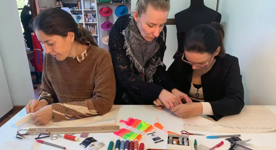 Tre donne si dedicano al cucito a mano, circondate da fili colorati, forbici e cartamodelli.
