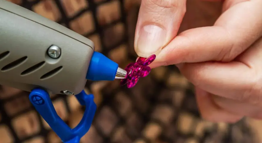Applicazione strass su tessuto con colla a caldo. Una mano tiene un fiorellino fucsia mentre la pistola a caldo è pronta per incollare.