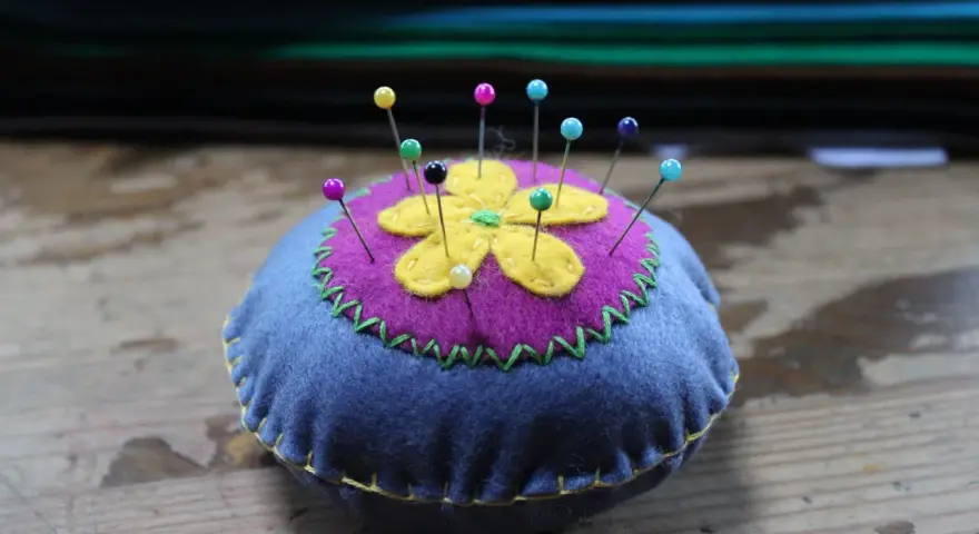 Puntaspilli fai da te a forma di fiore, con petali gialli su sfondo viola e base blu, decorato con cuciture a zig-zag verdi e gialle.