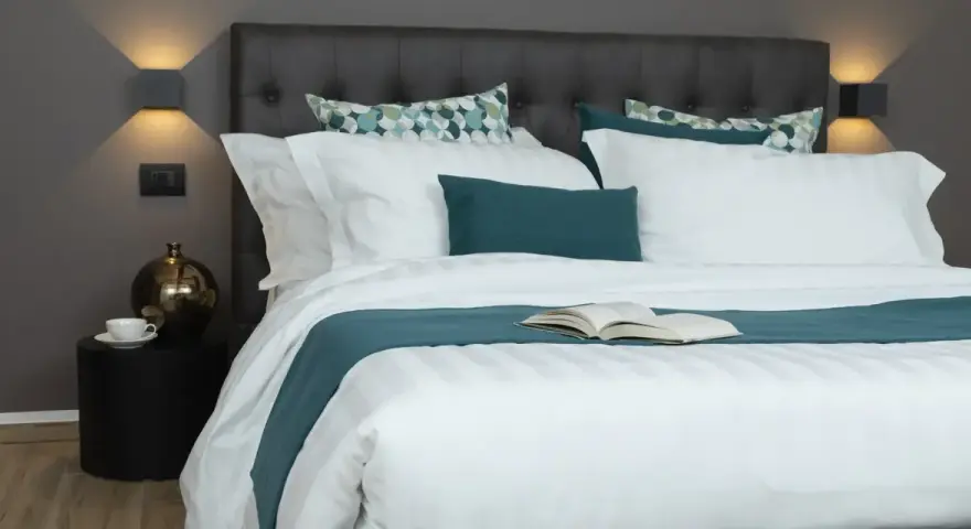 Letto con trapunta bianca a righe, cuscini coordinati e un libro aperto. Ecco come mettere la trapunta nel letto per un riposo perfetto.