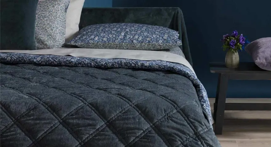 Letto con copriletto trapuntato blu scuro, cuscini floreali e un dettaglio che ricorda che il velluto si rovina con acqua.