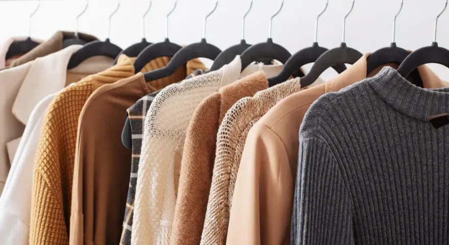 Una fila di capi d'abbigliamento in tonalità neutre, tra cui un maglione lavorato a maglia e un blazer in tessuto cupro lavaggio, pronti per essere indossati.