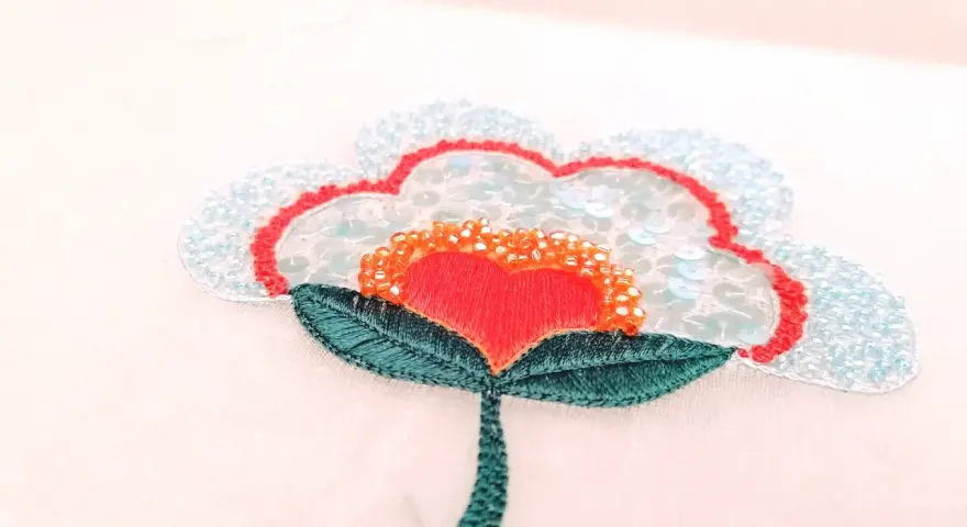 Un fiore stilizzato con petali azzurri e paillettes, un cuore rosso al centro e perline arancioni. Un'opera di ricamo francese che cattura la luce.