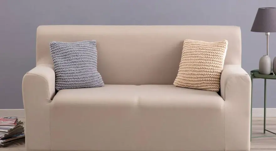 Divano con copridivano in tessuto color crema, due cuscini decorativi a maglia, uno grigio e uno beige.