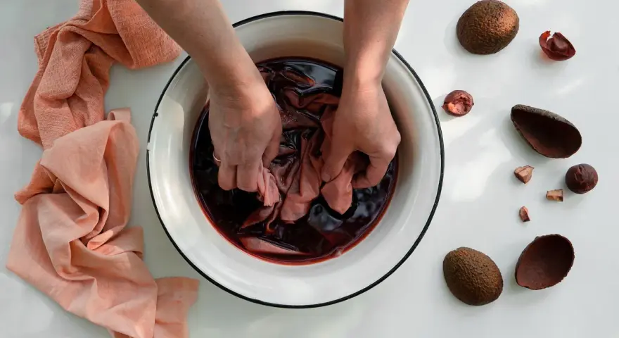 Mani che tingono tessuti in una bacinella con liquido scuro, circondata da bucce di avocado. Un processo naturale per come tingere i tessuti.