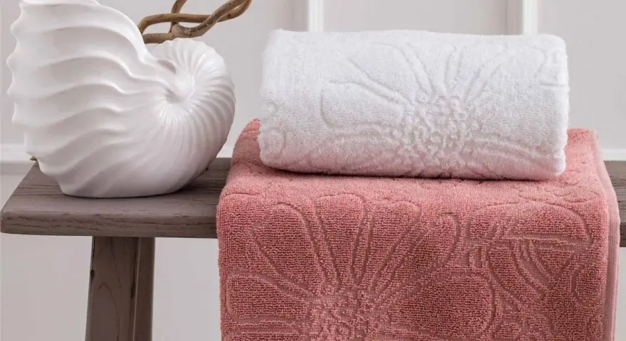 Asciugamani rosa e bianchi con motivi floreali in rilievo, disposti su un tavolino con una decorazione a conchiglia. La grammatura degli asciugamani suggerisce morbidezza e qualità.
