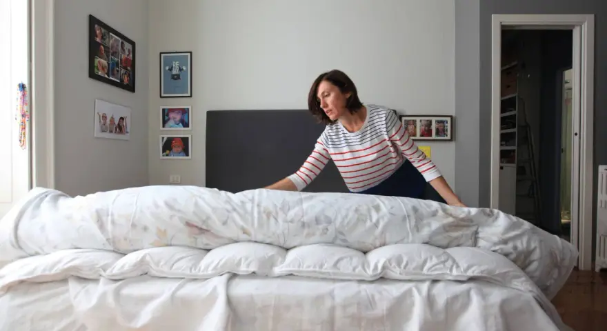Donna con maglia a righe prova a mettere il copripiumino su un letto disfatto.