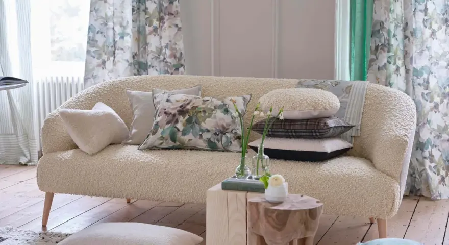 Divano in tessuto bouclé color crema, pronto per essere rifoderato, con cuscini decorativi e tende floreali sullo sfondo.