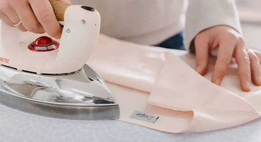 Mani che stirano lenzuola rosa pallido con un ferro da stiro Immetec. Un'etichetta con il logo "Daffodil" è visibile sul tessuto.