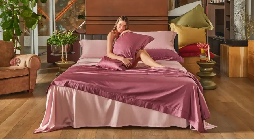Donna sorridente su un letto con lenzuola rosa. Scopri come lavare le lenzuola nuove per mantenerle morbide e fresche.