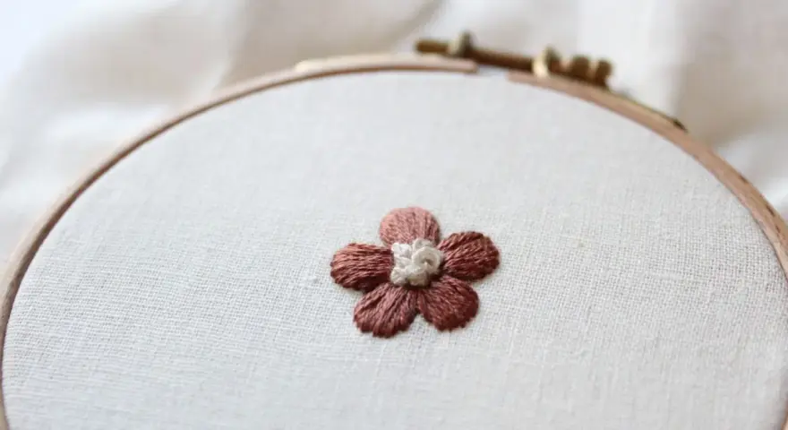 Piccolo fiore ricamato a punto pieno su tela grezza, con petali marroni e centro bianco.