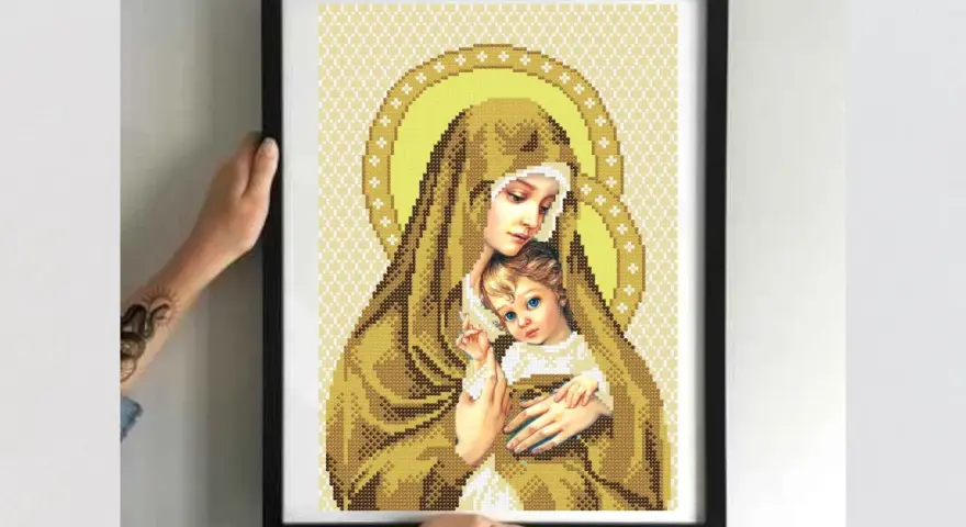Cross-stitch of Madonna and Child in a black frame, held by hands.