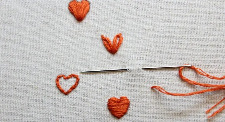 Un ago con filo arancione pronto per creare un altro cuore ricamato a mano su tela grezza.