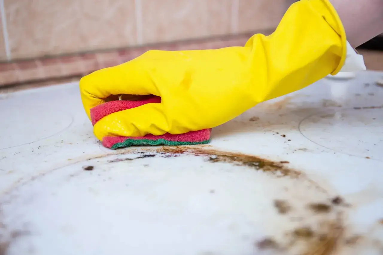 Mano con guanto giallo pulisce piano cottura con spugna. Scopri come togliere le macchie gialle dalla biancheria e dalla cucina.