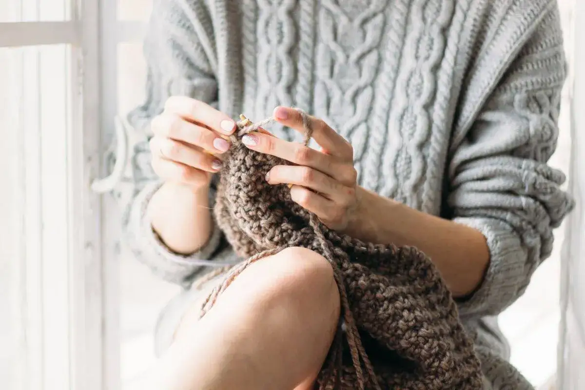 Mani che lavorano a maglia con filato marrone, indossando un maglione grigio a trecce. Un momento di tranquillit&agrave; per lavare lana a mano.