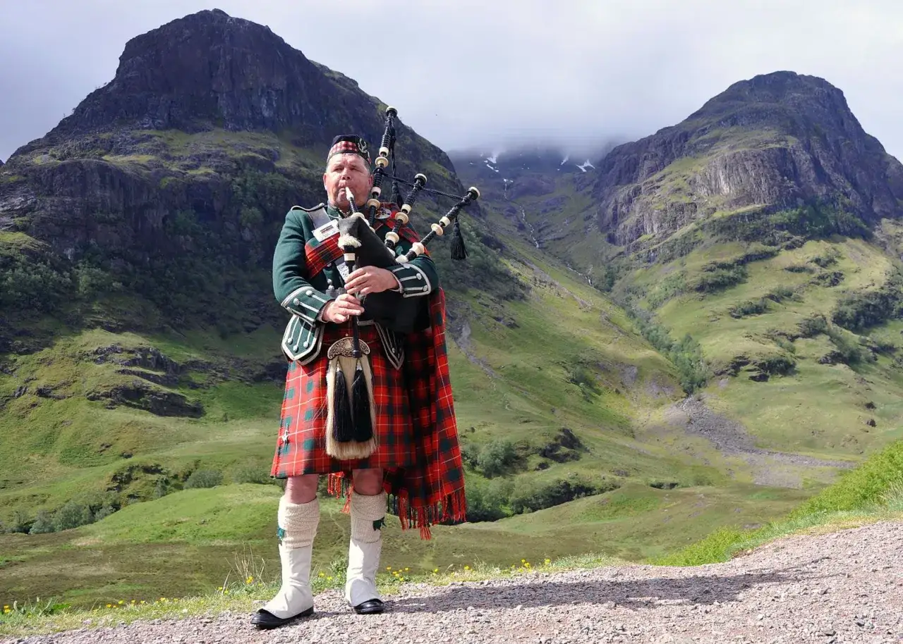 Uomo scozzese con cornamuse indossa un kilt a quadri rossi e verdi, un capo tradizionale che racconta storie di terre selvagge.
