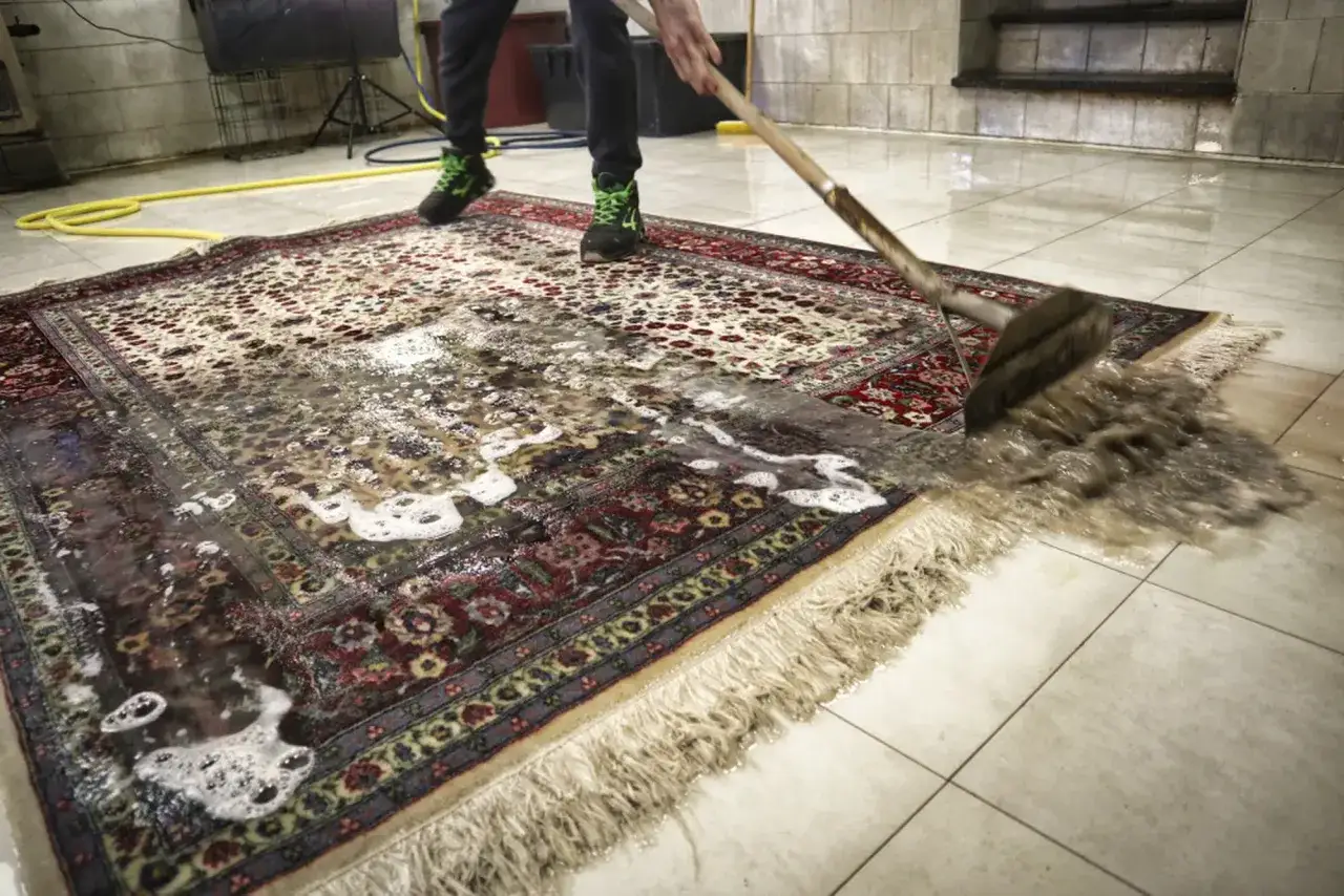 Persona con scarpe verdi lava un tappeto persiano con una scopa, rimuovendo acqua e sporco. Il processo di tappeti lavaggio è in corso.