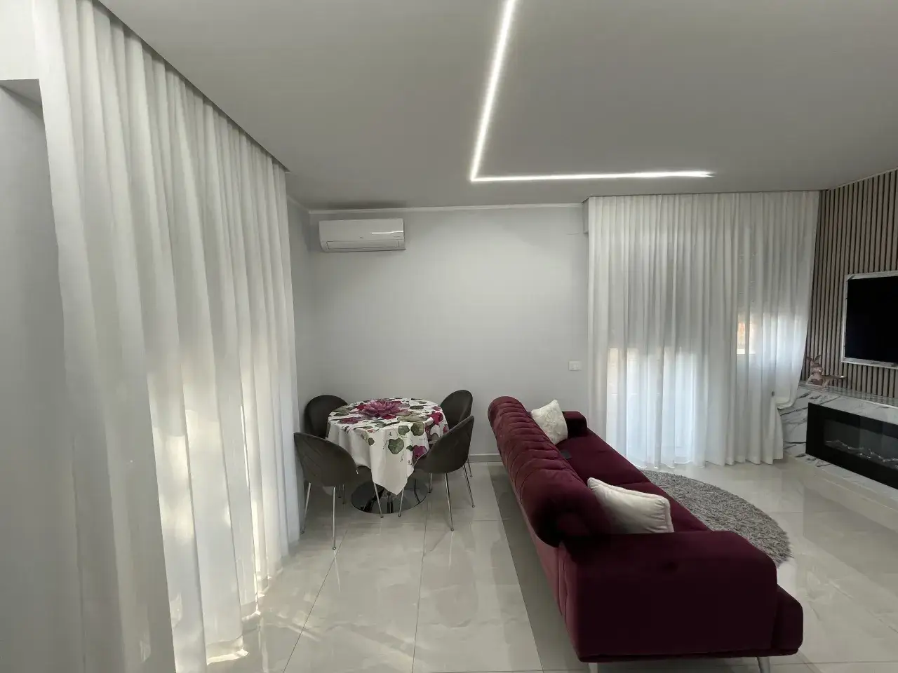 Tende bianche e leggere creano un'atmosfera intima, nascondendo l'esterno. Un divano bordeaux e un tavolo da pranzo completano l'arredo.