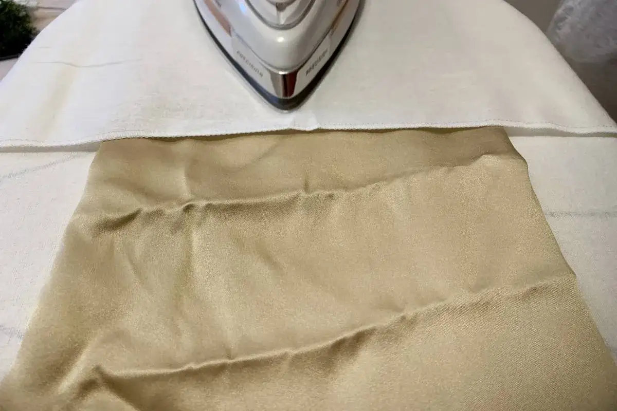 Un ferro da stiro su un asse, pronto a stirare un tessuto color champagne. Il raso si può stirare con cura per eliminare le pieghe.